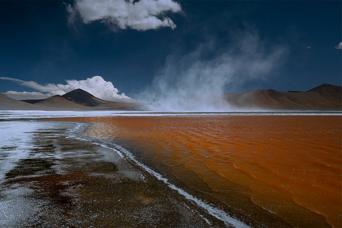 Steam rising off an orange lake with mountains beyond