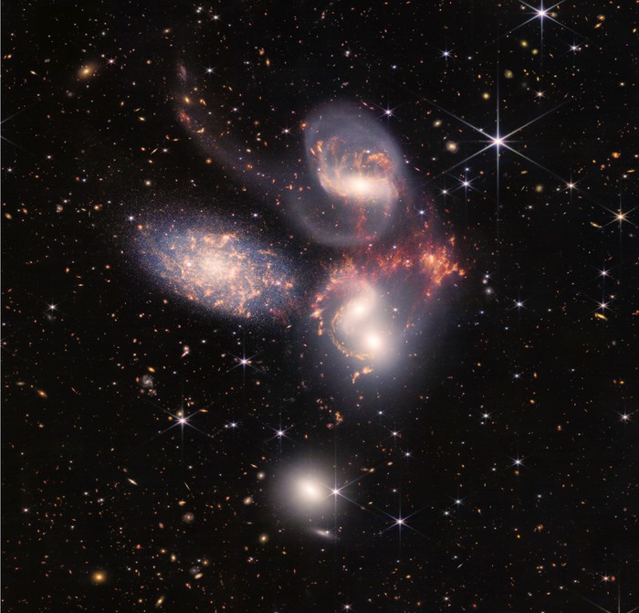A group of five galaxies that appear close to each other in the sky: two in the middle, one toward the top, one to the upper left, and one toward the bottom. Four of the five appear to be touching. One is somewhat separated. In the image, the galaxies are large relative to the hundreds of much smaller (more distant) galaxies in the background. All five galaxies have bright white cores. Each has a slightly different size, shape, structure, and coloring. Scattered across the image, in front of the galaxies are number of foreground stars with diffraction spikes: bright white points, each with eight bright lines radiating out from the center.